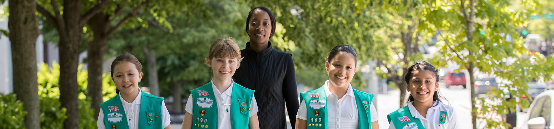  Adult volunteer troop leader with junior Girl Scout troop in uniform vests and sashes walking outside. 